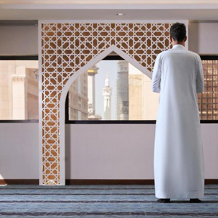 Makarem Ajyad Makkah Hotel Мека Екстериор снимка The photo depicts a person standing with their back facing the viewer, wearing a traditional white garment. They are in a spacious interior setting that features an ornate archway with intricate patterns, typical of Islamic architecture. Through the 