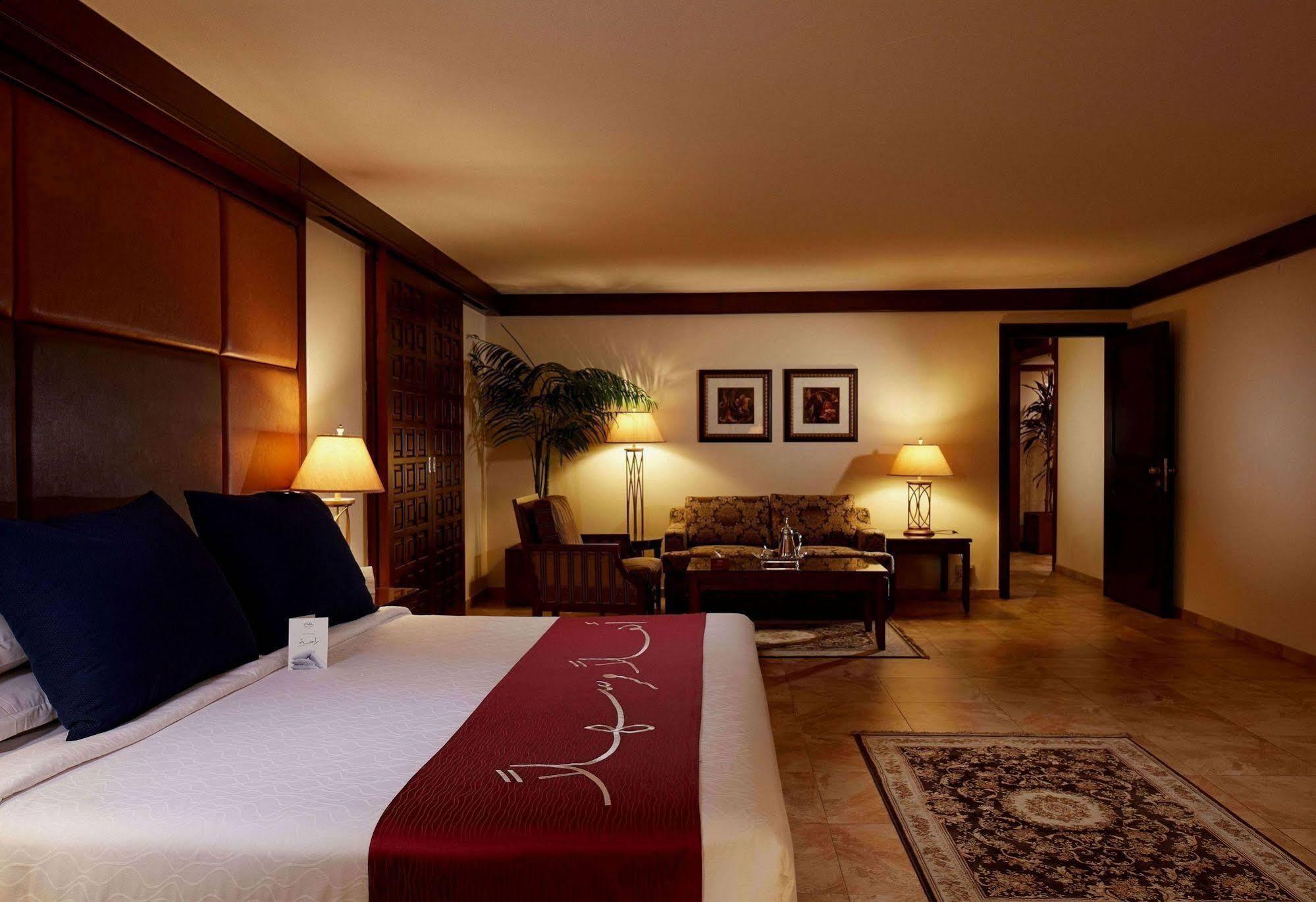 Makarem Ajyad Makkah Hotel Мека Екстериор снимка The photo depicts a cozy hotel room with a warm and inviting atmosphere. It features a large bed with dark blue pillows and a maroon throw blanket. On either side of the bed, there are lamps providing soft lighting. In the background, a small seating