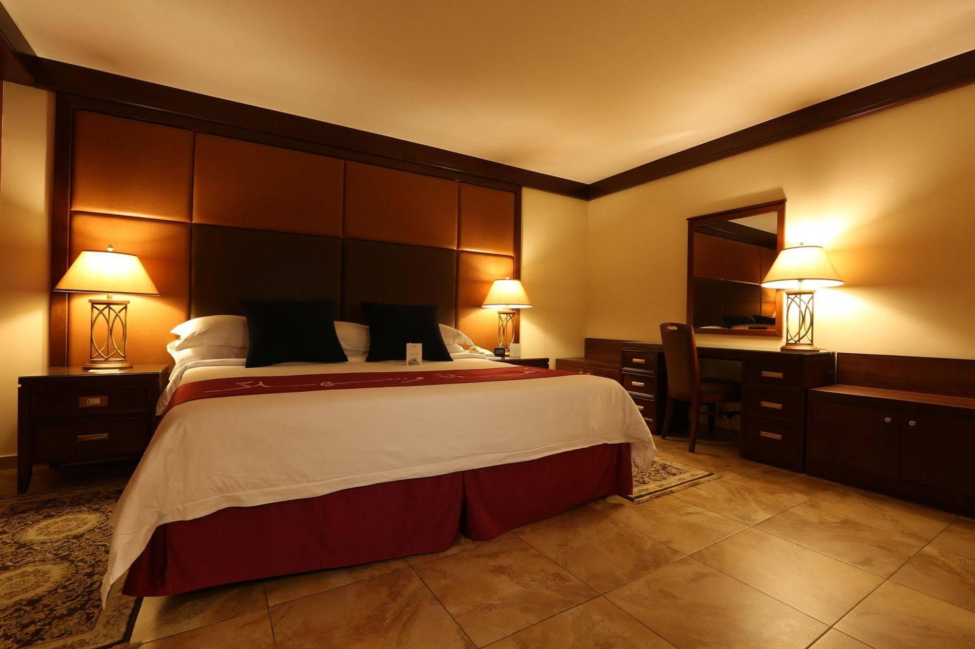 Makarem Ajyad Makkah Hotel Мека Екстериор снимка The photo shows a well-appointed hotel room with a cozy and inviting atmosphere. It features a large bed with a white comforter and dark pillows, positioned against a wooden headboard. There are two bedside lamps providing soft lighting on either sid