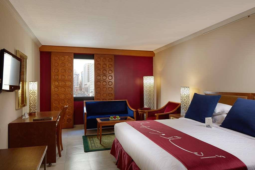 Makarem Ajyad Makkah Hotel Мека Стая снимка The photo showcases a hotel room featuring a modern design. In the center, there's a large bed with white linens and a decorative red runner. On either side of the bed, there are wooden nightstands with lamps. A blue sitting area is visible, composed