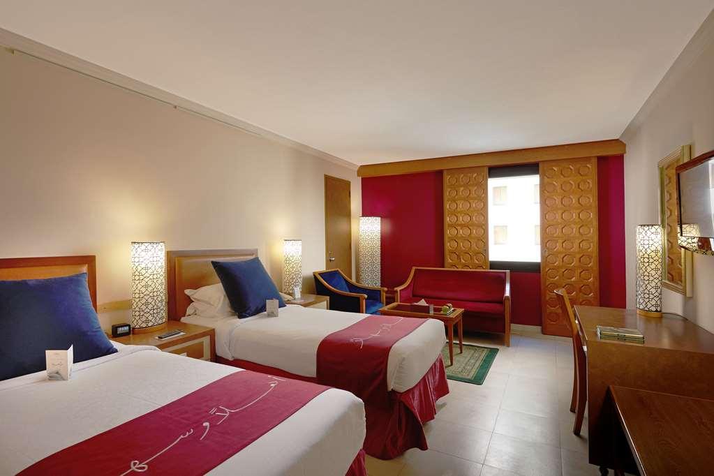 Makarem Ajyad Makkah Hotel Мека Стая снимка The photo shows a hotel room featuring two twin beds with red and white bedding. There are decorative lamps on either side of the beds. A small seating area is visible, including a blue chair and a red sofa. There is a wooden desk against one wall, a