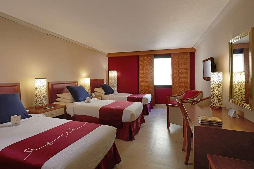 Makarem Ajyad Makkah Hotel Мека Стая снимка The photo shows a hotel room featuring three beds arranged with red and white bedding. The walls are painted in a neutral tone, and there are decorative lamps on nightstands beside each bed, providing ambient lighting. One of the walls has a window c