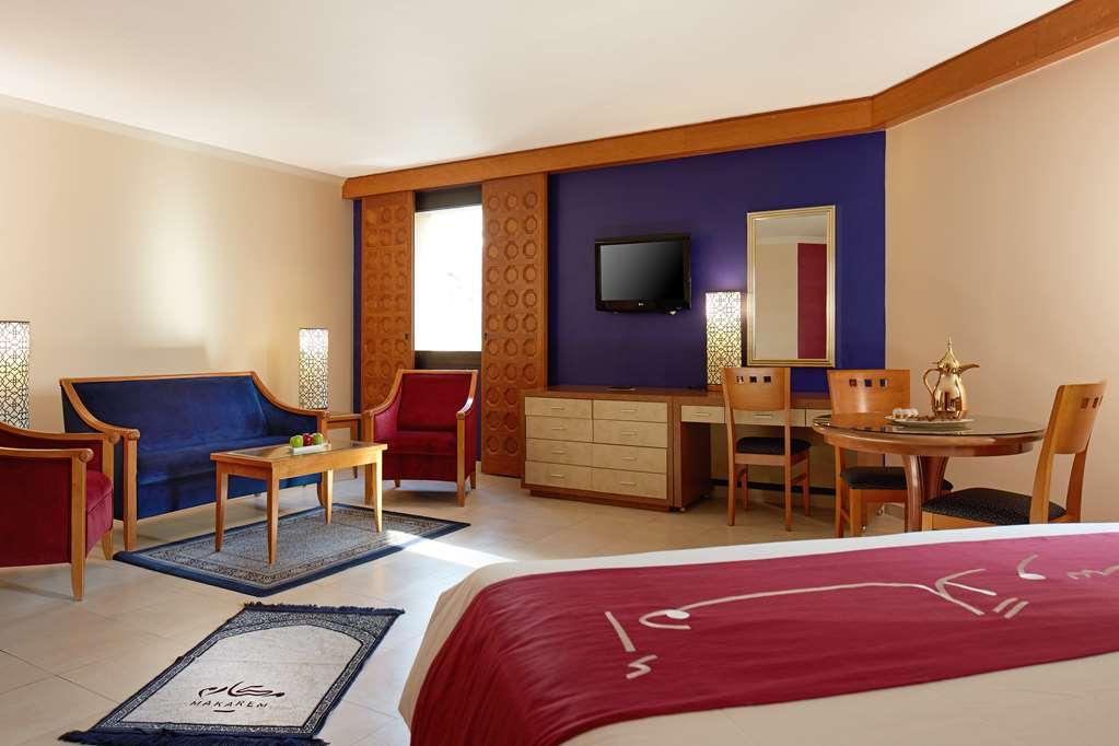 Makarem Ajyad Makkah Hotel Мека Стая снимка The image shows a hotel room with a modern, comfortable design. The room features a large bed with a red and white bedspread, a seating area that includes a blue sofa, two red chairs, and a wooden coffee table. There's a flat-screen TV mounted on the