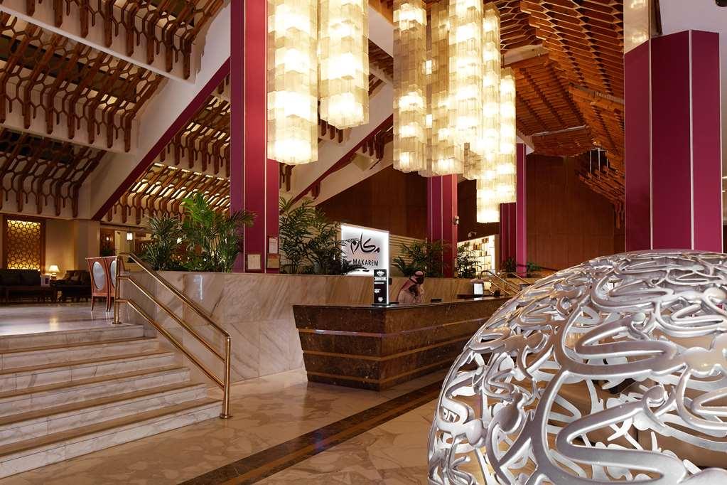 Makarem Ajyad Makkah Hotel Мека Интериор снимка The photo depicts a luxurious hotel lobby. It features a grand entrance with a staircase leading up to a reception area. The lobby has high ceilings adorned with striking, modern light fixtures that hang down, providing a warm ambiance. The walls are