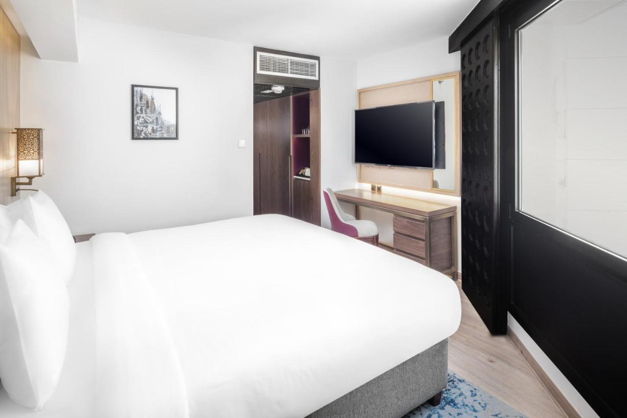 Makarem Ajyad Makkah Hotel Мека Екстериор снимка The photo shows a modern hotel room featuring a large bed with white linens at the forefront. In the background, there is a wall-mounted television across from a desk with a chair. A wardrobe or cabinet is located to the side, and there's a decorativ