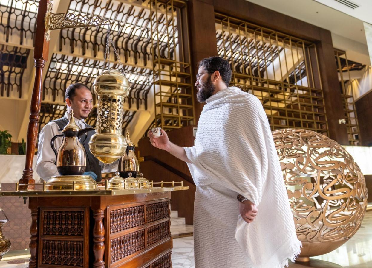 Makarem Ajyad Makkah Hotel Мека Екстериор снимка The photo depicts a scene in a luxurious setting, likely a hotel or a fine dining establishment. In the foreground, a man wearing traditional attire, possibly a white garment, is engaging with a staff member. The staff member is behind a wooden count