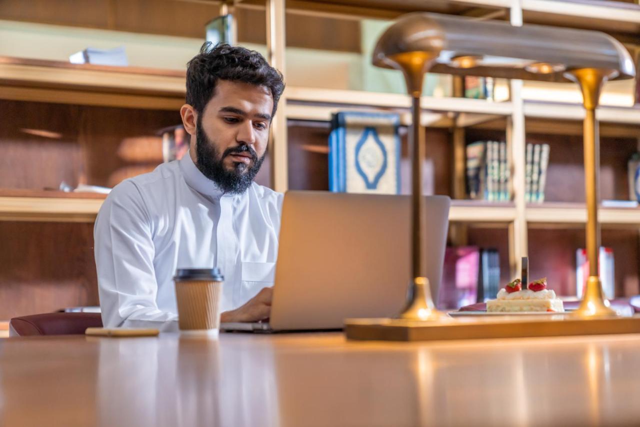 Makarem Ajyad Makkah Hotel Мека Екстериор снимка The photo shows a man sitting at a desk in a library or a study area. He is wearing a white traditional garment and is focused on working on a laptop. In front of him, there is a cup of coffee and a small cake decorated with strawberries. The backgro