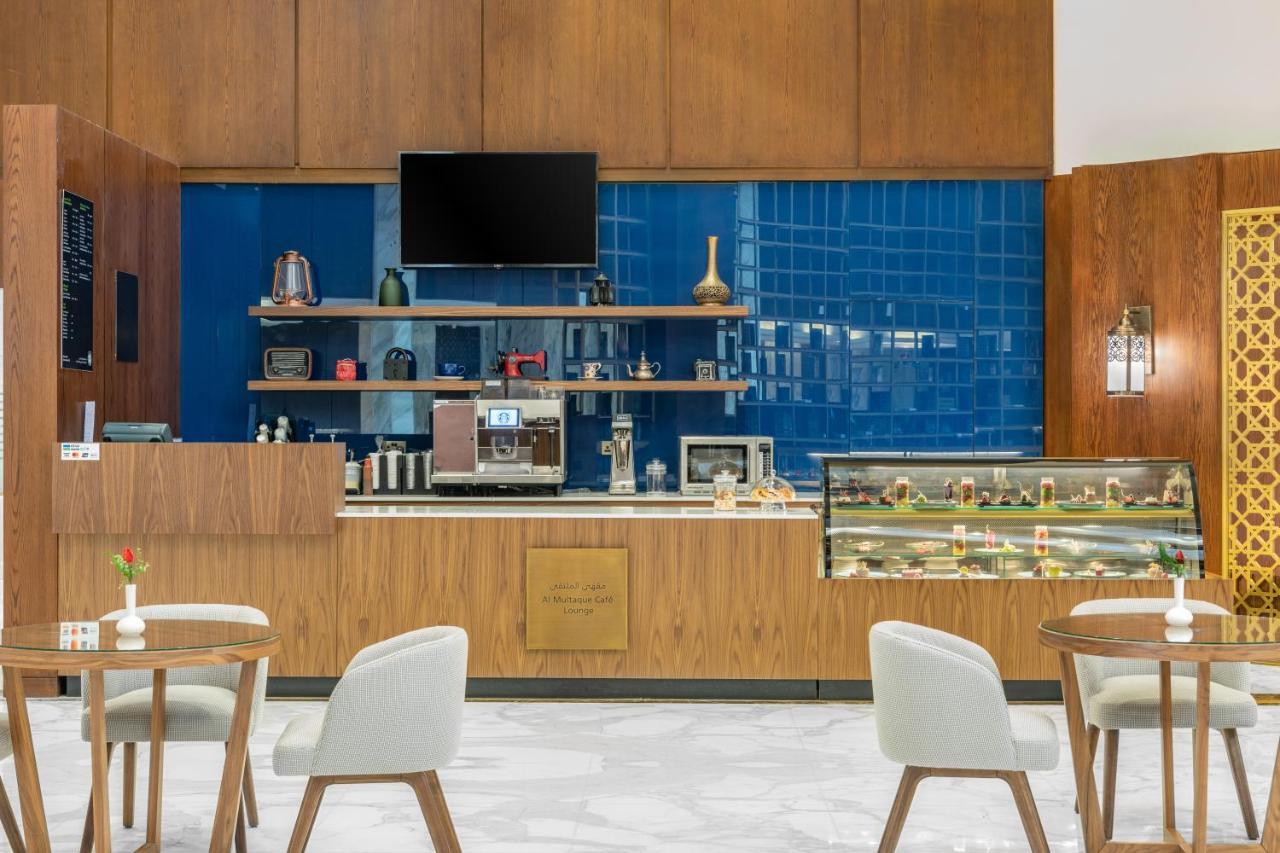 Makarem Ajyad Makkah Hotel Мека Екстериор снимка The photo shows the interior of a café. The design features warm wood paneling and a striking blue tiled wall. In the foreground, there are several light-colored chairs and small tables arranged neatly. Behind the counter, there's a variety of coffee