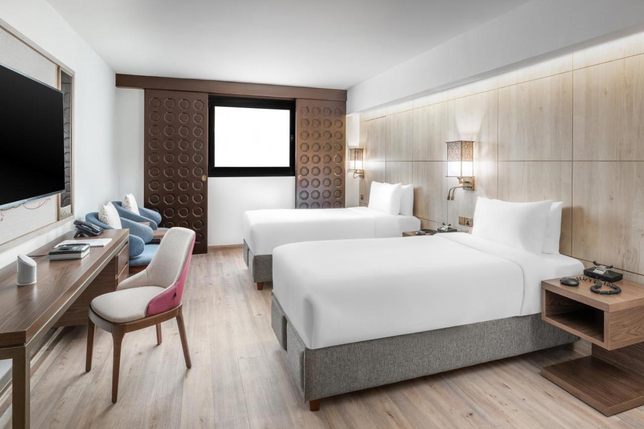 Makarem Ajyad Makkah Hotel Мека Екстериор снимка The photo shows a modern hotel room featuring two beds with white linens. There is a wooden accent wall with a window above it. Opposite the beds, a wall-mounted television is visible. A stylish wooden desk with a chair is situated on one side of the