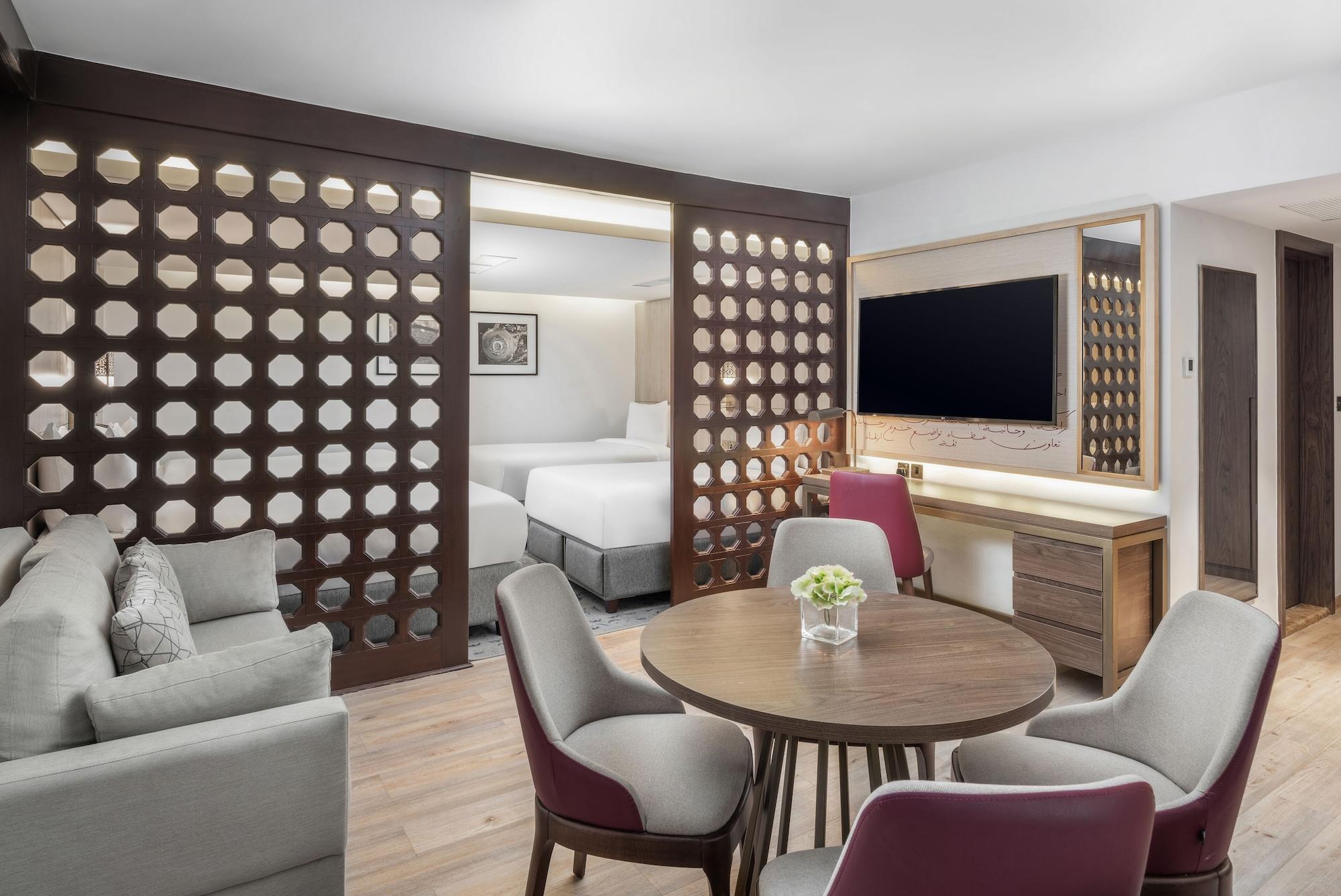 Makarem Ajyad Makkah Hotel Мека Екстериор снимка The photo shows a modern hotel room or suite. It features a living area with a comfortable gray sofa and a round wooden dining table surrounded by upholstered chairs. A large flat-screen TV is mounted on the wall opposite the seating area. There's a 