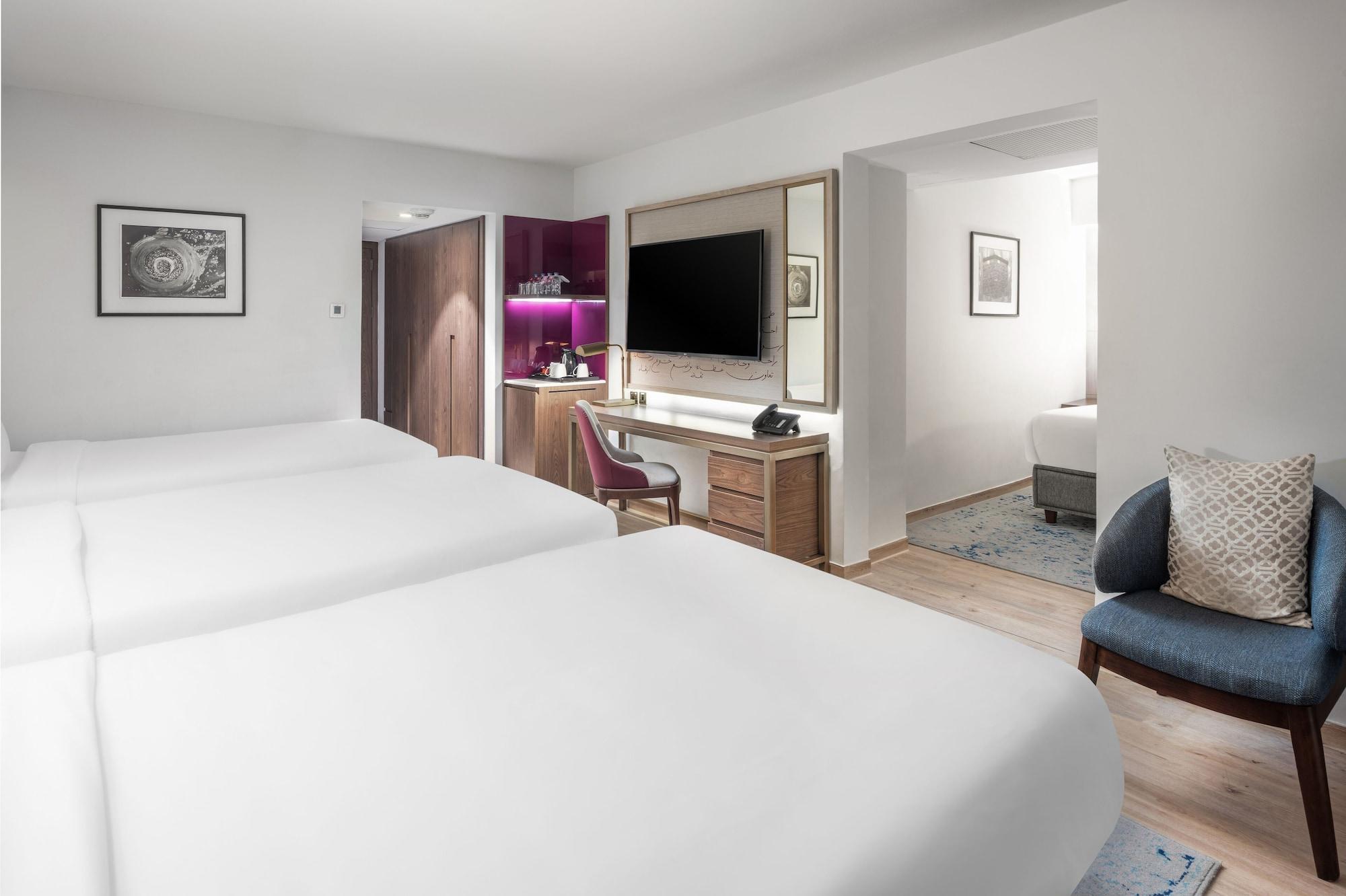 Makarem Ajyad Makkah Hotel Мека Екстериор снимка The photo shows a modern hotel room featuring two white beds with smooth linens. The room has a minimalistic design with light-colored walls and wooden flooring. On one side, there is a desk and chair next to a television mounted on the wall, along w