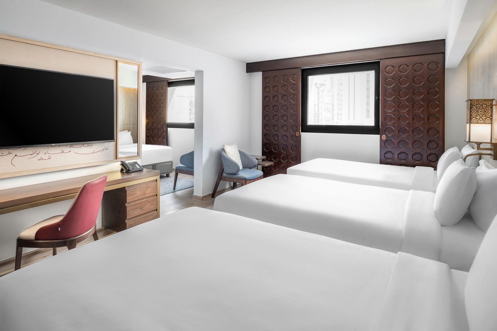 Makarem Ajyad Makkah Hotel Мека Екстериор снимка The photo shows a hotel room featuring three beds with white linens. There is a large flat-screen TV mounted on the wall opposite the beds, and a wooden side table beneath it. A red chair is positioned near the TV, while a blue upholstered chair is p