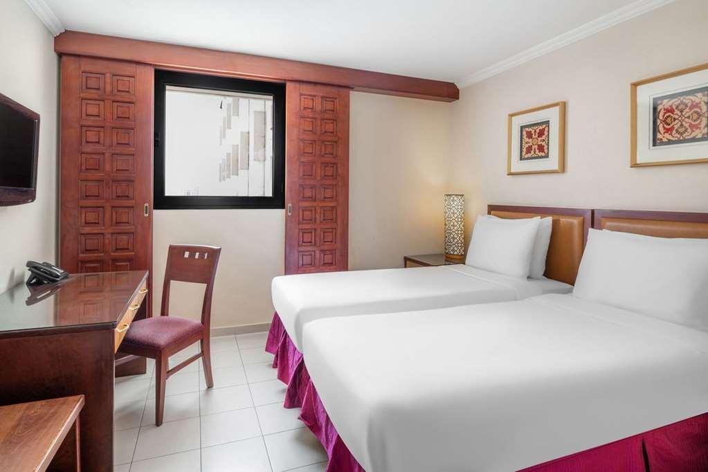 Makarem Ajyad Makkah Hotel Мека Стая снимка The photo shows a hotel room with two beds, each covered with white bedding. There is a wooden desk and a chair near the beds. The room features a window with a wooden frame and a decorative lamp beside the beds. On the wall, there are two framed art