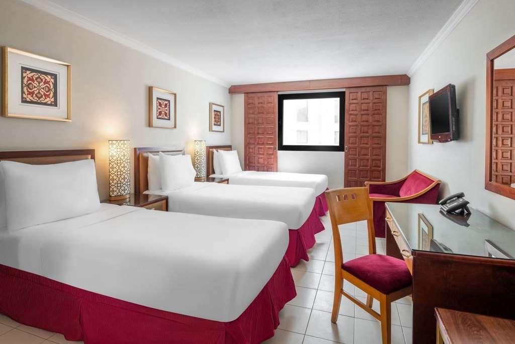 Makarem Ajyad Makkah Hotel Мека Стая снимка The photo shows a hotel room featuring two twin beds with white linens and red bed skirts. There are decorative elements on the walls, including framed artwork. A large window provides natural light, while a desk and a chair are positioned next to it