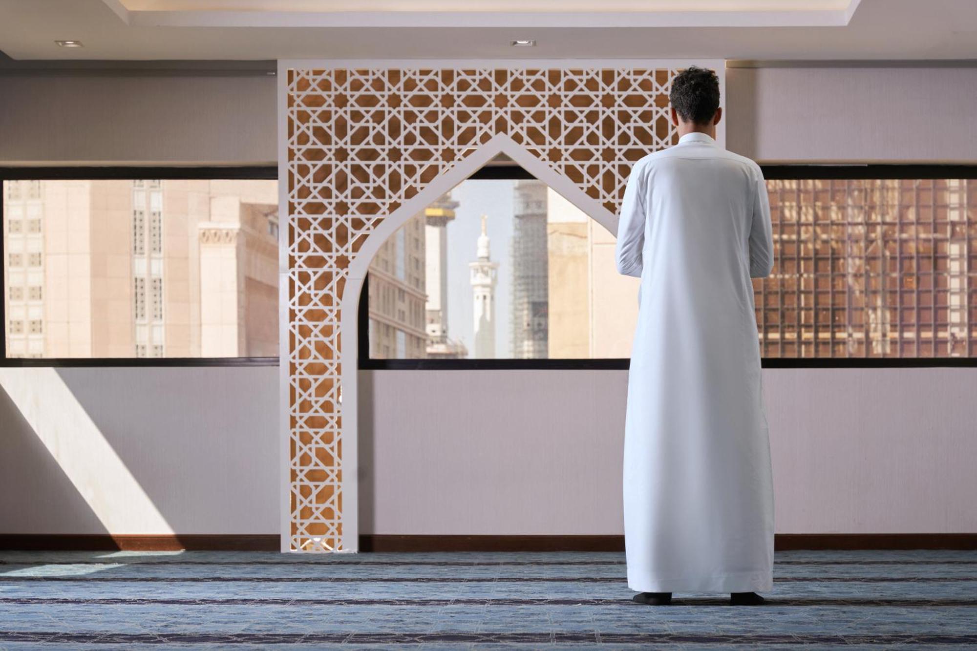 Makarem Ajyad Makkah Hotel Мека Екстериор снимка The photo depicts a person standing with their back facing the viewer, wearing a traditional white garment. They are in a spacious interior setting that features an ornate archway with intricate patterns, typical of Islamic architecture. Through the 