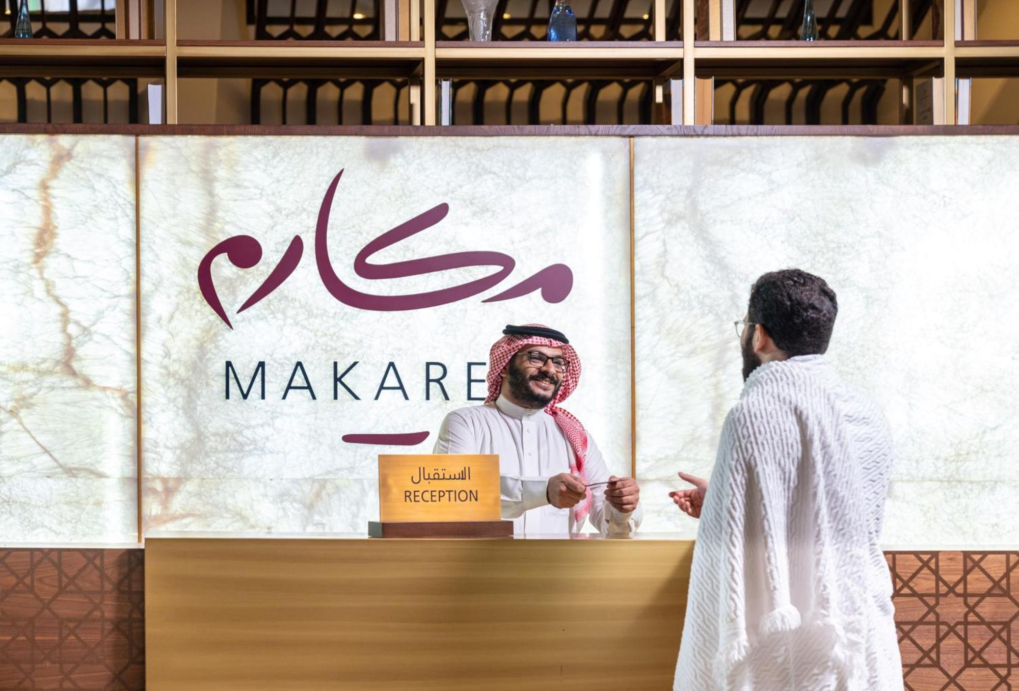 Makarem Ajyad Makkah Hotel Мека Екстериор снимка The photo depicts a reception area in a hotel or establishment named "Makarem." In the image, a receptionist, dressed in traditional attire, is smiling and interacting with a guest who is wearing a white robe, commonly known as a thobe or ihram. The 