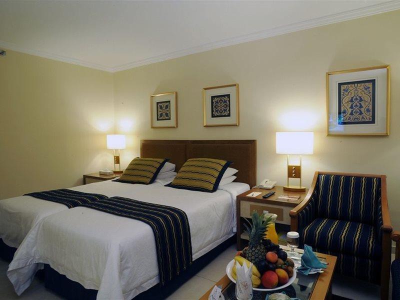 Makarem Ajyad Makkah Hotel Мека Екстериор снимка The photo shows a hotel room with a cozy and inviting atmosphere. It features two beds with white linens and dark striped bedspreads. There are two bedside lamps providing warm light, and the walls are decorated with framed artworks. Beside the beds 