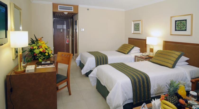 Makarem Ajyad Makkah Hotel Мека Екстериор снимка The photo shows an interior of a hotel room featuring two twin beds with white linens and green decorative accents. The walls are painted in a light beige color, and there is a small table next to one of the beds. A desk with a chair is positioned ne