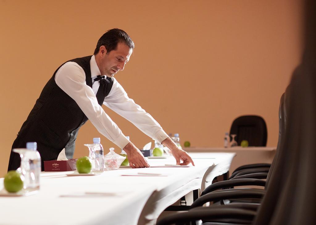 Makarem Ajyad Makkah Hotel Мека Екстериор снимка The image shows a waiter or service staff member in a formal attire, which includes a vest and bow tie. He is preparing a long table, likely for an event or meeting. The table is set with items such as water bottles and green apples, and there are se