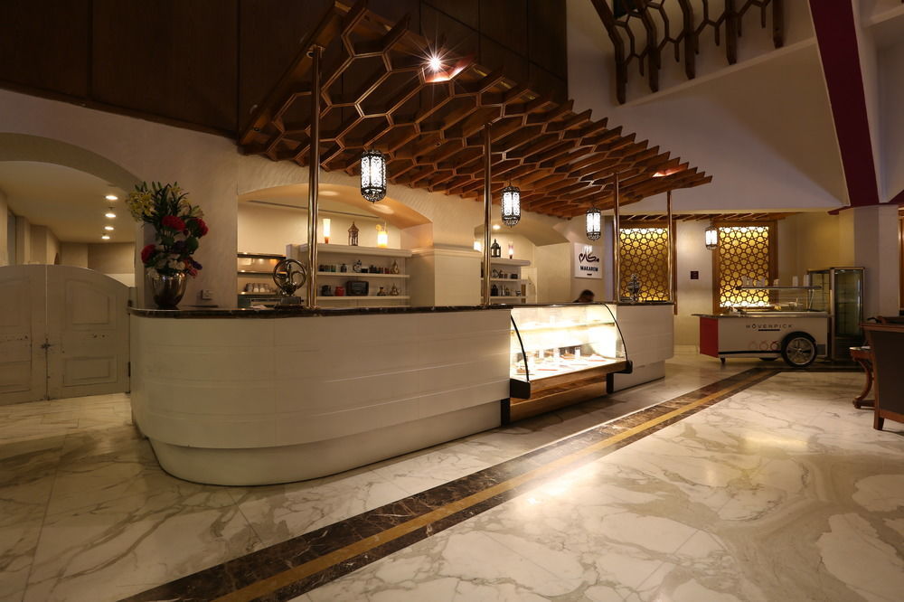 Makarem Ajyad Makkah Hotel Мека Екстериор снимка The photo shows a modern reception area or lobby of a hotel or restaurant. The focal point is a curved front desk made of white material, featuring a glass display case that showcases various items. Above the desk, there is a decorative wooden struct
